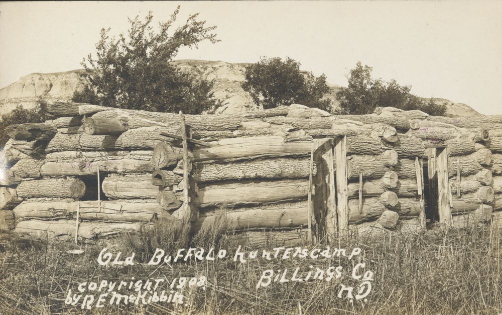 The roofless log walls of an abandoned cabin in long grass below badland bluffs, with two empty doorways.