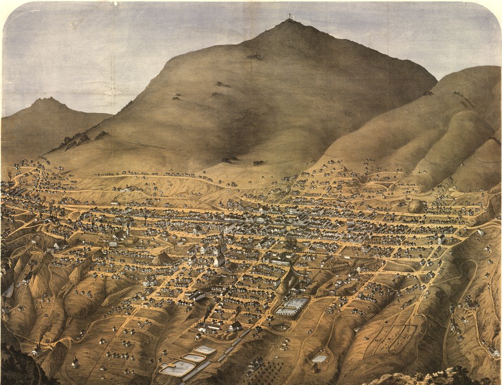 A coloured bird's-eye drawing of Virginia City laid out in a grid of streets on the flank of a large bare mountain, with mills, reservoirs and mine works. The surrounding hills are brown and almost treeless.
