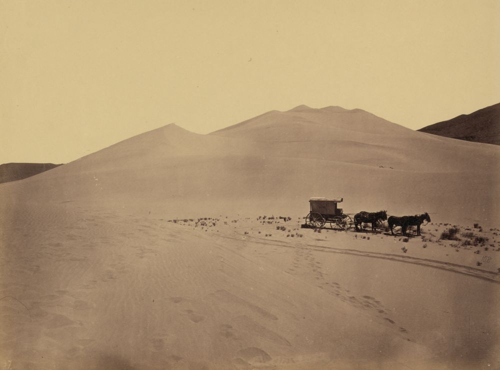 Smooth, high sand dunes under a pale sky, with a small covered wagon and its two-pair mule team standing on the flat sand in front, and wheel tracks and footprints in the foreground.