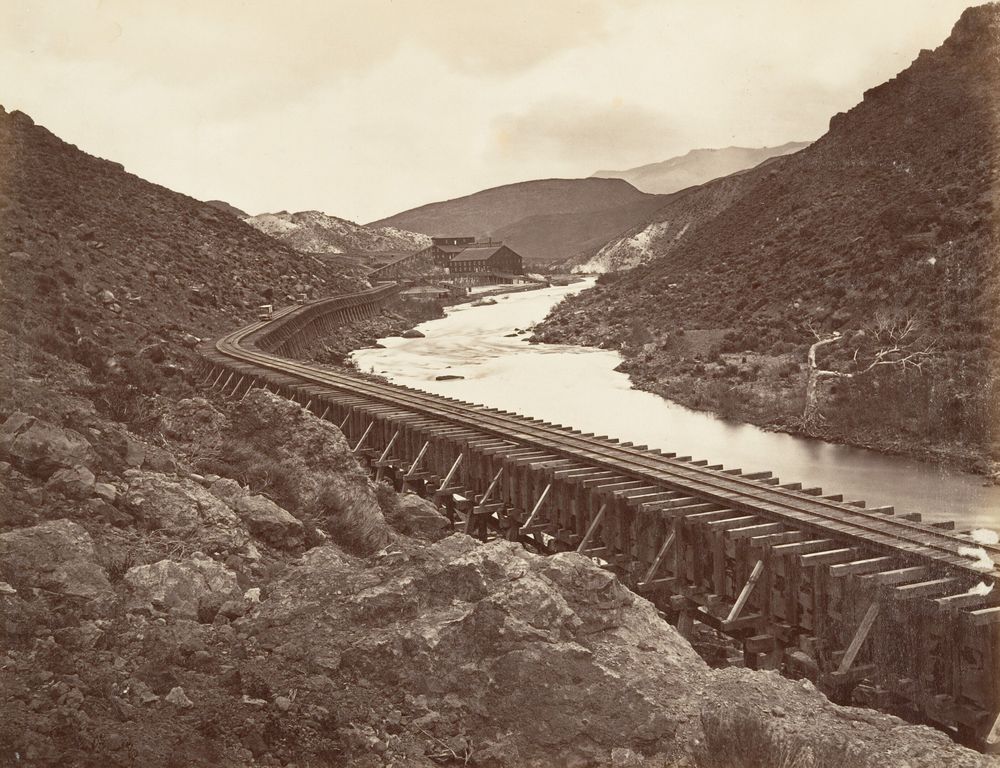 A long wooden flume on trestles running along a river down a rocky, almost treeless canyon toward a mill building in the distance.