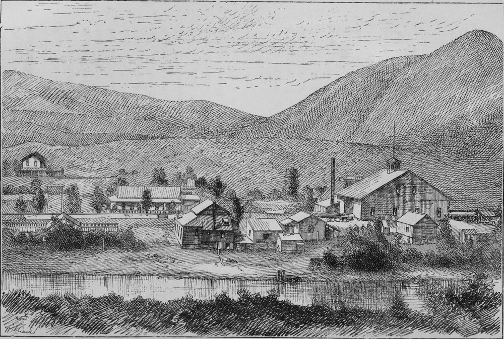 A cluster of mill buildings with a tall chimney beside a river, below steep bare hills.