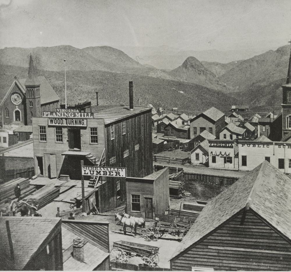 Rooftops of a wooden town looking down a canyon to bare hills. In the middle, a planing mill with a Lumber sign and a yard stacked with sawn boards, a horse team and wagon in the street.