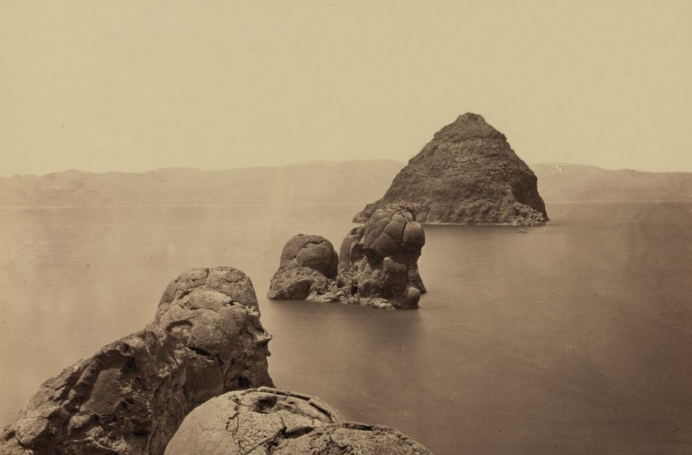 A pyramid-shaped rock island rising from a wide, still lake, with rounded rock domes in the middle distance and large weathered boulders in the foreground.