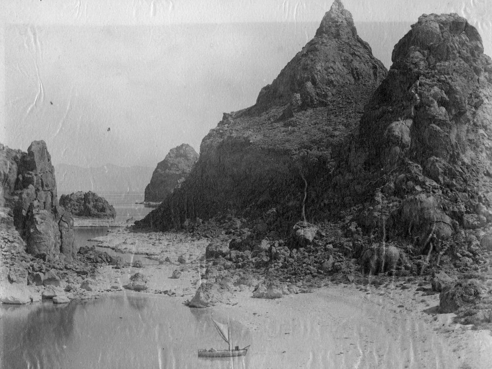 Tall, jagged rock spires rising above a shallow bay and a rock-strewn beach, with a small sailing boat on the still water.