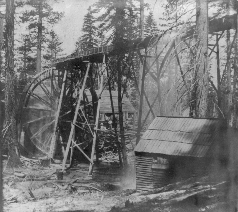 A large wooden water wheel and a raised flume on timber trestles among tall pine trees, with a small shingled shed in front and sawn logs and slabs on the ground.