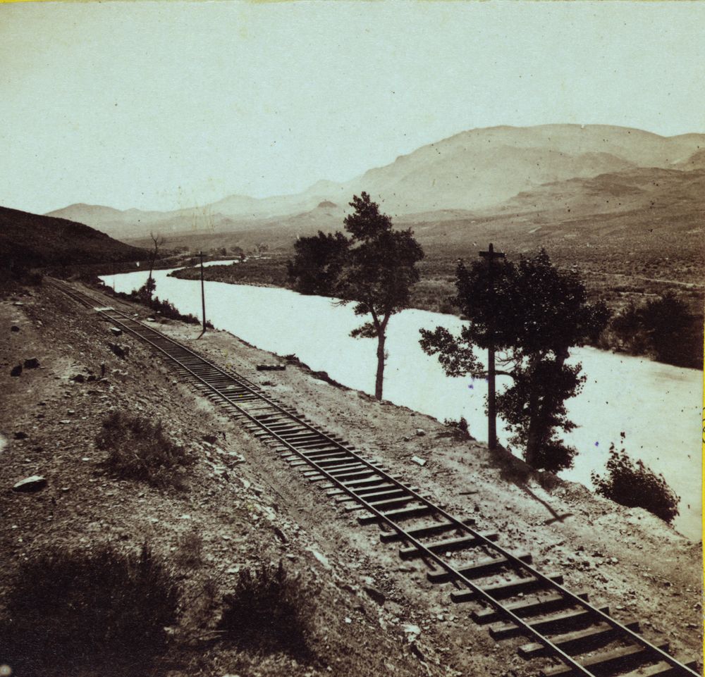 A wide, pale river curving through a dry valley toward a bare mountain, with two pine trees on its bank and a railroad track running along the near shore through sage and stones.