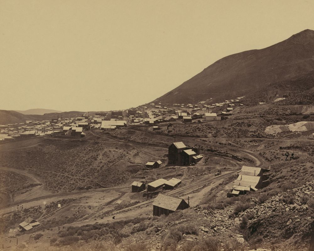 A town of wooden houses strung along the foot of a bare mountain slope. In the foreground, mine buildings, sheds and a curving rail grade, with pale waste dumps on the right. No trees on any of the hillsides.