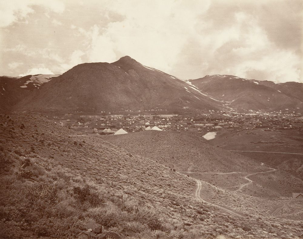 A bare, dark mountain under heavy cloud, with a spread of wooden buildings and pale heaps of mine waste at its foot. In the foreground, sage-covered slopes with wagon roads winding across them and almost no trees.